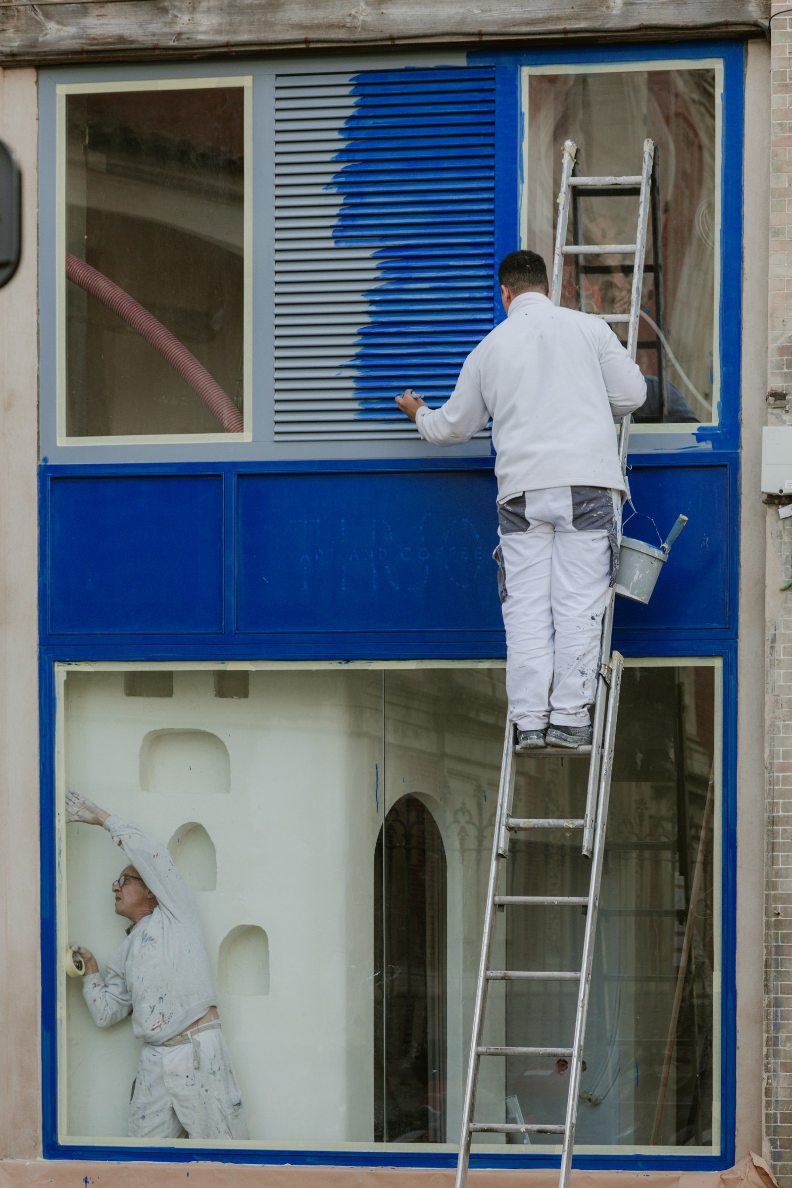 Painter in a white uniform on a ladder, rolling deep blue paint onto a building facade